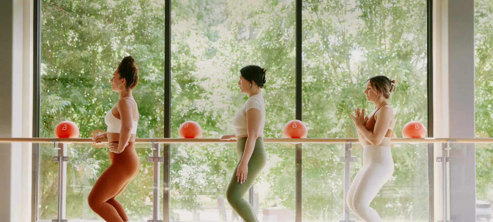 3 women working at a barre in profile against a backdrop of a wall of windows and trees