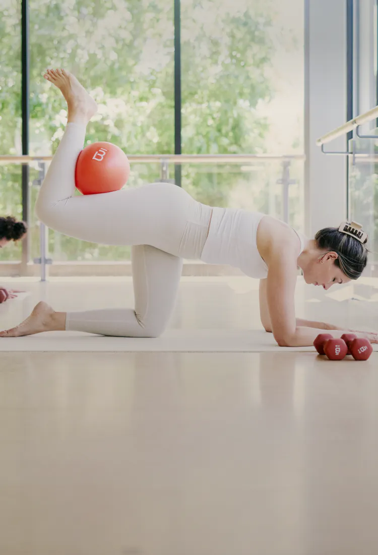 3 women working on their backs on a mat in profile against a backdrop of a wall of windows and trees